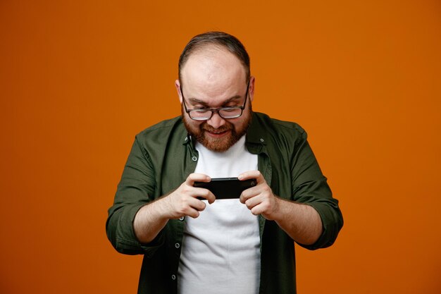 Bebaarde man in vrijetijdskleding met een bril die een smartphone vasthoudt en naar het scherm kijkt terwijl hij spelletjes speelt, blij en opgewonden over oranje achtergrond