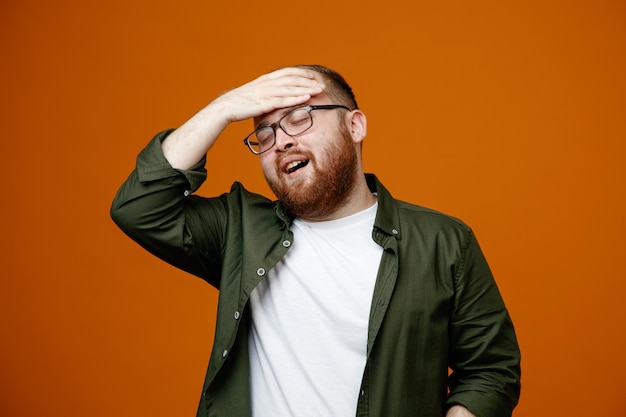 Bebaarde man in vrijetijdskleding met een bril die er verward uitziet terwijl hij de hand op zijn voorhoofd houdt en over een oranje achtergrond staat