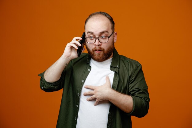Bebaarde man in vrijetijdskleding met een bril die verbaasd kijkt terwijl hij op een mobiele telefoon praat die over een oranje achtergrond staat