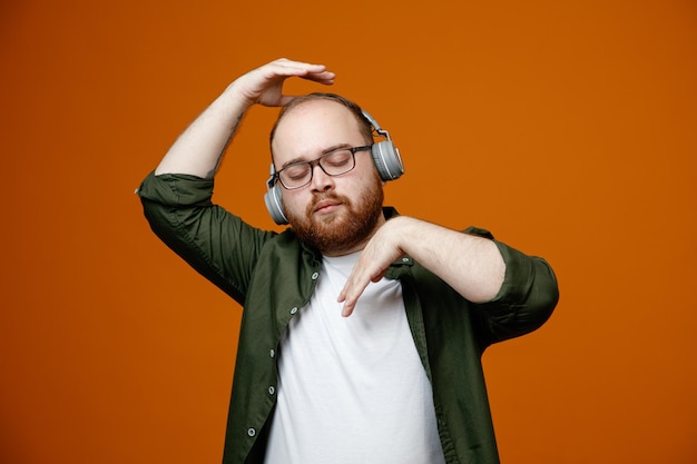 Gratis foto bebaarde man in vrijetijdskleding met een bril met een koptelefoon, kalm en ontspannen luisterend naar muziek die over een oranje achtergrond staat