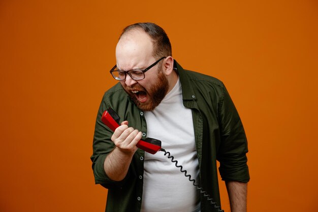 Bebaarde man in vrijetijdskleding met een bril met een retro-telefoon die schreeuwt met een agressieve uitdrukking die over een oranje achtergrond staat