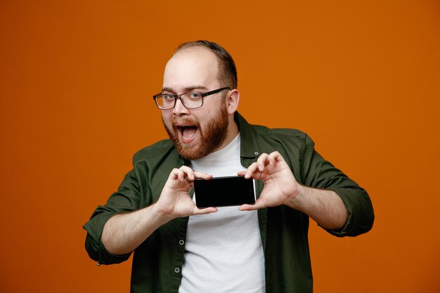 Bebaarde man in vrijetijdskleding met een bril waarop een smartphone te zien is die blij en positief glimlacht naar de camera terwijl hij over een oranje achtergrond staat