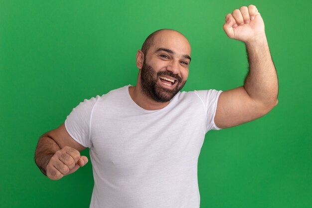 Bebaarde man in wit t-shirt blij en opgewonden het opheffen van vuisten die zich over groene muur bevinden