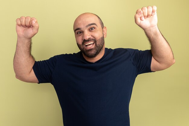 Bebaarde man in zwart t-shirt gek, blij en opgewonden schreeuwen, gebalde vuisten verheugd over zijn succes staande over groene muur