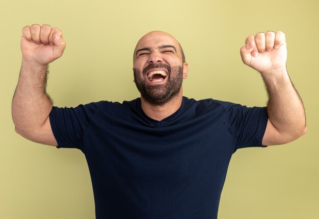 Bebaarde man in zwart t-shirt gek, blij en opgewonden schreeuwende gebalde vuisten staande over groene muur