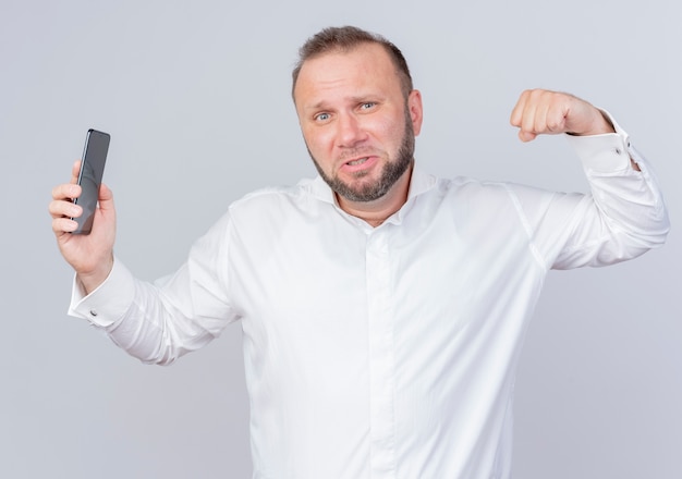 Bebaarde man met een wit overhemd dat smartphone toont met balde vuist die biceps toont die zelfverzekerd over witte muur staan