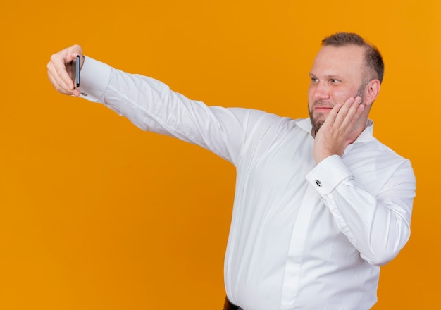 Bebaarde man met een wit overhemd doet selfie lachend met blij gezicht staande over oranje muur