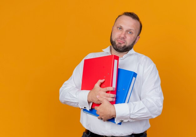 Bebaarde man met een wit overhemd met mappen verward met droevige uitdrukking staande over oranje muur