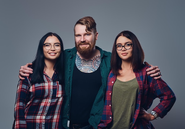 Bebaarde man met tatoeages op zijn lichaam en twee brunette hipster vrouwen geïsoleerd over grijze achtergrond.