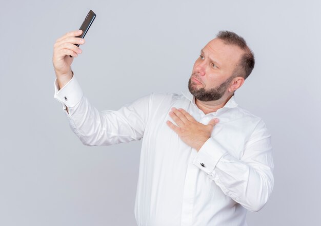 Bebaarde man met wit overhemd met smartphone met videogesprek dankbaar gevoel hand op borst staande over witte muur