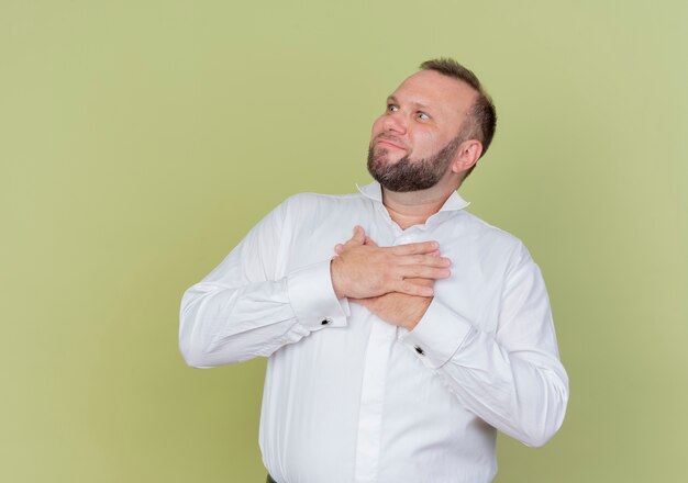 Bebaarde man met wit overhemd opzij kijken hand in hand op de borst dankbaar gevoel staande over lichte muur