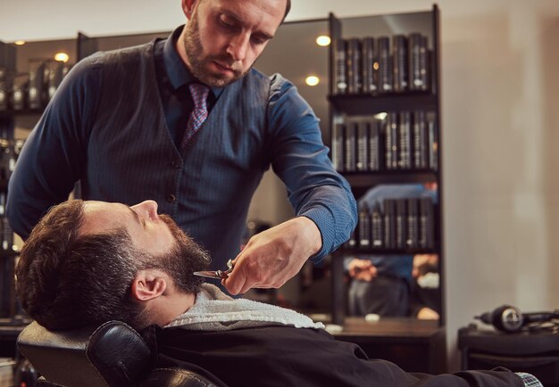 Bebaarde man zittend in een fauteuil in een kapperszaak terwijl kapper baard modelleren met een schaar en kam bij de kapperszaak.