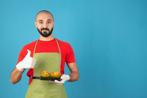 Bebaarde mannelijke chef-kok in schort en handschoenen met een donkere houten plank met gebak.