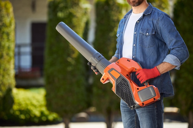 Gratis foto bebaarde mannelijke tuinman in handschoenen met krachtige draadloze bladblazer buitenshuis gewasweergave van klusjesman