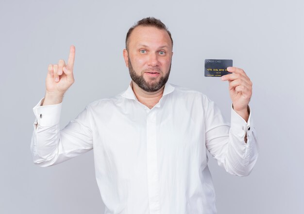 Bebaarde mens die wit overhemd draagt dat creditcard toont die wijsvinger toont die met gelukkig gezicht glimlacht met nieuw idee dat zich over witte muur bevindt