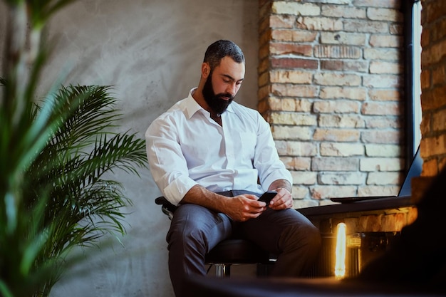 Bebaarde moderne man met smartphone in een kamer met loft interieur.