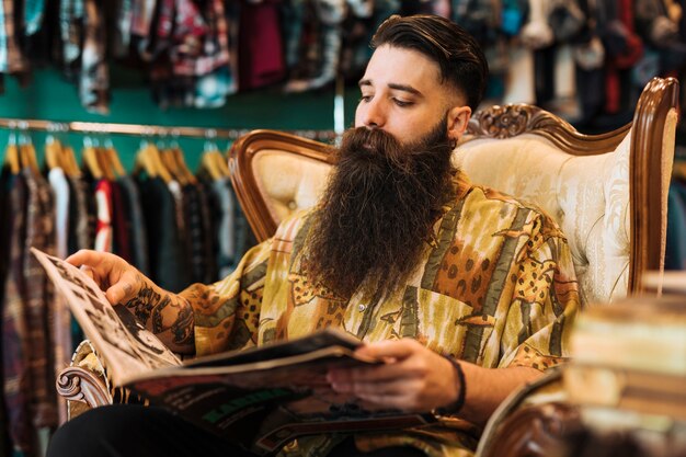 Bebaarde modieuze jonge mensenzitting op stoel die tijdschrift in de winkel bekijken