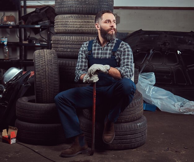 Bebaarde monteur gekleed in een uniform, houdt een koevoet vast, zit op oude autobanden in de garage.