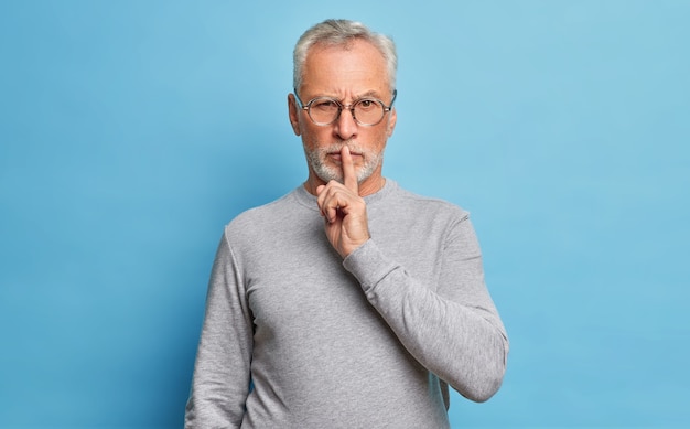 Bebaarde senior man heeft ernstige uitdrukking maakt stil gebaar vraagt stil te zijn met vinger op lippen eist stilte draagt optische bril en trui met lange mouwen geïsoleerd op blauwe muur