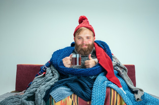 Bebaarde zieke man met rookkanaal zittend op de bank thuis of studio met kopje thee bedekt met warme gebreide kleding. ziekte, influenza-concept. ontspanning thuis. gezondheidszorgconcepten.