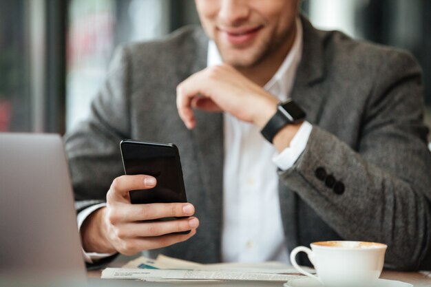 Bebouwd beeld van gelukkige zakenmanzitting door de lijst in koffie en het gebruiken van smartphone