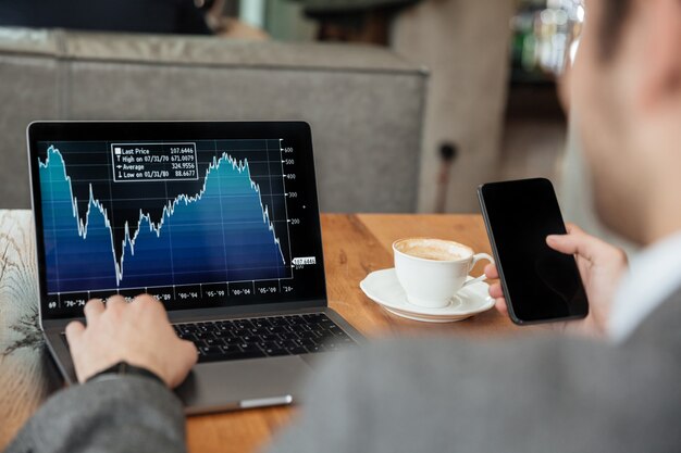 Bebouwd beeld van zakenmanzitting door de lijst in koffie en het analyseren van indicatoren op laptop computer terwijl het gebruiken van smartphone