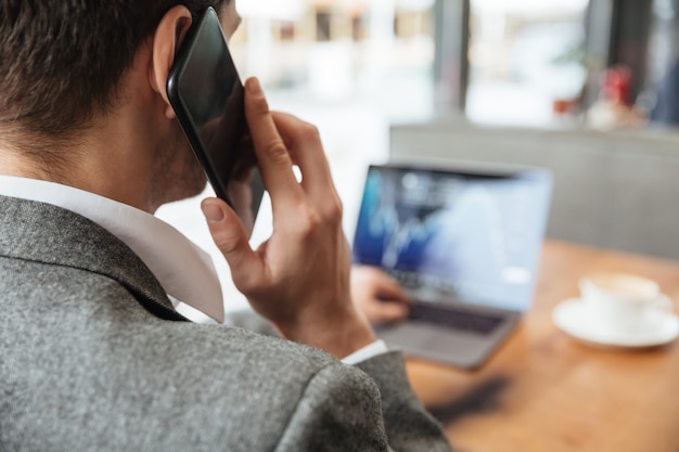 Bebouwd beeld van zakenmanzitting door de lijst in koffie en het analyseren van indicatoren op laptop computer terwijl het spreken door smartphone. Focus op man