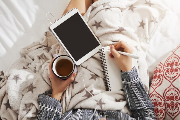 Bed is mijn kantoor. Hoge hoek schot van meisje liggend in gezellige sfeer, muziek luisteren in oortelefoons, tablet te houden en thee te drinken terwijl het maken van aantekeningen in haar notitieblok. Creatief meisje probeert nieuw gedicht te maken