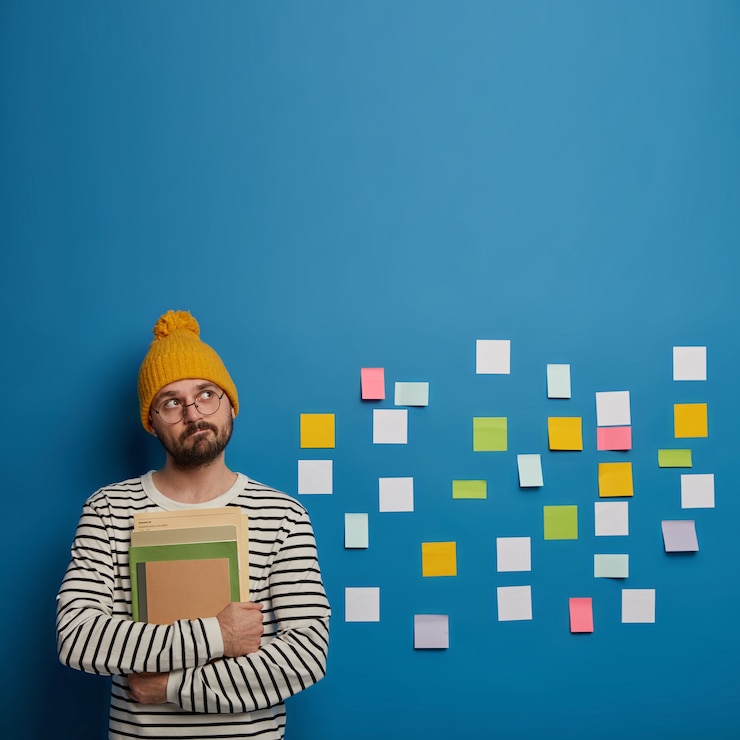 Gratis Foto bedachtzame man met baard houdt boeken vast, kijkt peinzend naar boven, denkt na over hoe hij een project kan maken, brainstormt over verschillende ideeën in gedachten