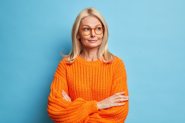 bedachtzame schattige blonde veertig jaar oude tevreden vrouw houdt armen over elkaar denkt aan iets en kijkt weg draagt een bril gebreide trui.