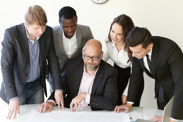 Bedrijfs brainstorm en werk onder een groot project bij ontwerpbureau. Vier collega's in kantoorkleren staan aandachtig naar de plannen te kijken. Tekeningen lagen op de tafel naast hun hoofdleider.