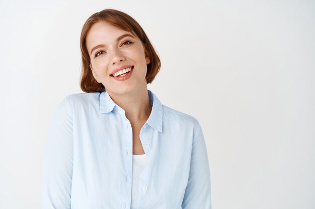 Bedrijfs- en mensenconcept. Portret van een glimlachende jonge vrouw in blouse die er gelukkig uitziet, staande op een witte muur