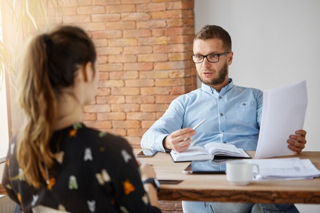 Bedrijfsconcept. Rijpe bebaarde ernstige mannelijke bedrijfsdirecteur die in bureau voor donkerharig meisje zit dat op sollicitatiegesprek kwam. Door papieren bladeren, praten over werkervaring.