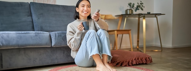 Gratis foto beeld van een jonge koreaanse vrouw die koffie drinkt, op afstand houdt van tv terwijl ze thuis televisie kijkt
