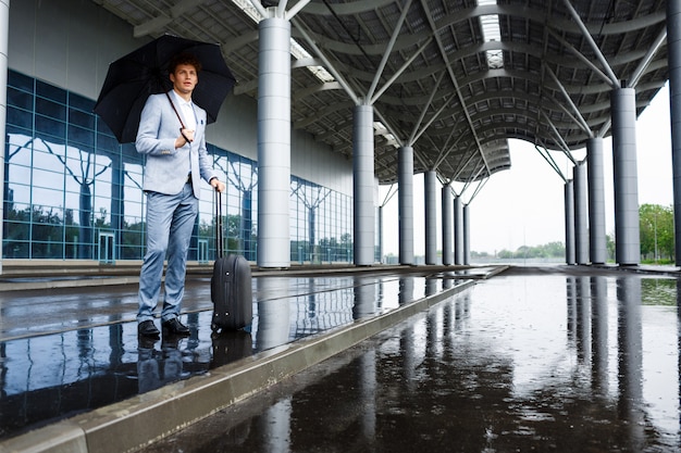 Beeld van jonge redhaired zakenman die zwarte paraplu in regen houden bij terminal