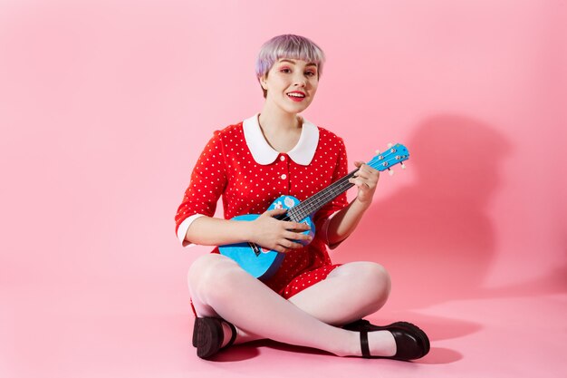 Beeld van mooi popachtig meisje met kort licht violet haar dat rode kleding draagt die blauwe ukelele over roze muur speelt