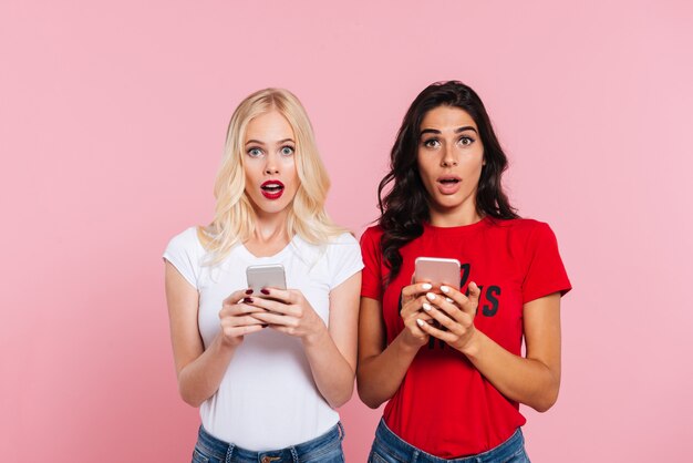Beeld van Twee behoorlijk geschokte vrouwen die smartphones houden en de camera over roze bekijken