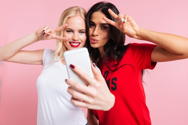 Beeld van twee mooie glimlachende vrouwen die selfie op smartphone over roze maken