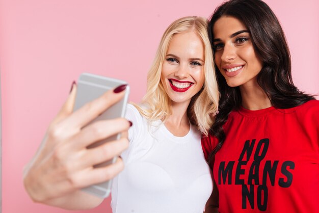 Beeld van twee vrij gelukkige vrouwen die selfie op smartphone over roze maken