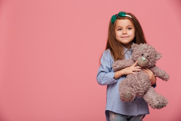 Beeld van zoet schoolmeisje met lang kastanjebruin haar dat houdend haar mooie teddybeer glimlacht