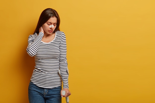 Beenblessures en beperking van menselijke bewegingen. Ontevreden vrouw raakt nek aan, voelt pijn, raakt gewond tijdens gevaarlijk reizen, heeft een gebroken neus, poseert tegen gele muur, lege ruimte opzij.