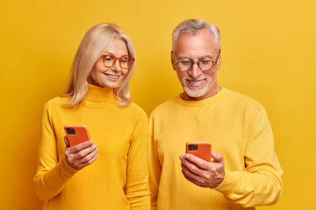bejaarde grootmoeder en grootvader bekijken samen foto's op smartphoneapparaten bekijk interessante grappige video online gekleed in casual gele coltruien poseren binnen
