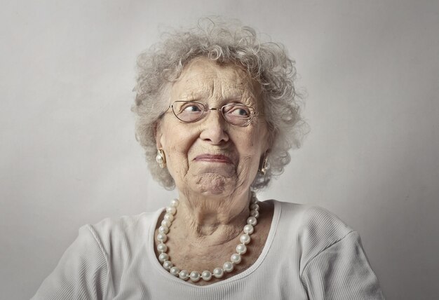 Bejaarde vrouw tegen een witte muur met een bezorgde blik op haar gezicht