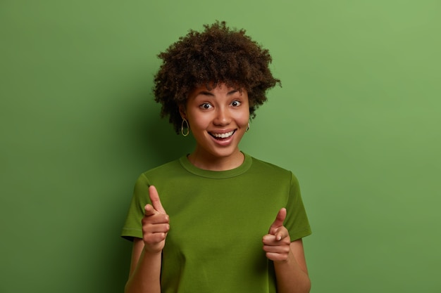 Gratis foto bekijken. vriendelijke positieve jonge afro-amerikaanse vrouw wijst wijsvinger naar je, kiest een persoon en nodigt uit om lid te worden van het team, feliciteert of prijst mooie baan, geïsoleerd over groene muur
