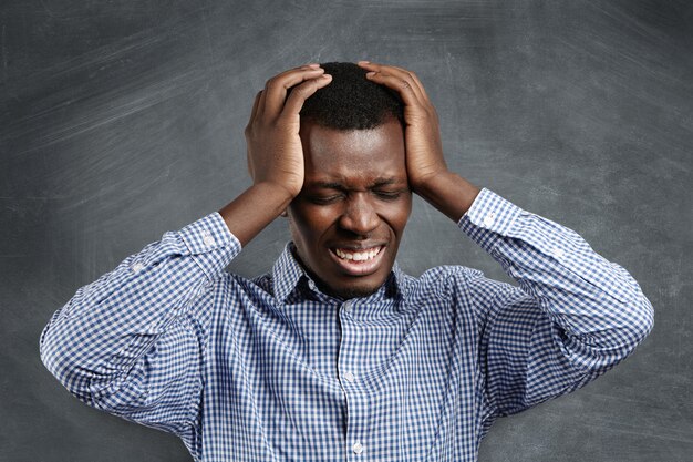 Beklemtoonde Afrikaanse zakenman die slechte hoofdpijn heeft, zijn hoofd drukt, ogen sluit en tanden klemt met pijnlijke gefrustreerde uitdrukking. Ondernemer met een donkere huidskleur die lijdt aan migraine