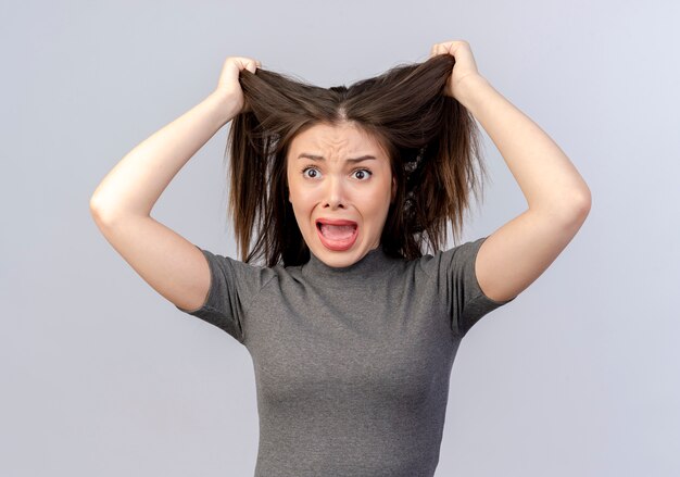 Beklemtoonde jonge mooie vrouw die recht kijkt die haar haar trekt dat op witte achtergrond wordt geïsoleerd