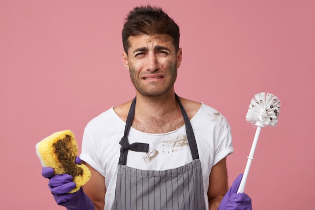 beklemtoonde trieste knappe student die bij roze muur staat met toiletborstel en spons jankend omdat hij huishoudelijk werk haat maar moet schoonmaken