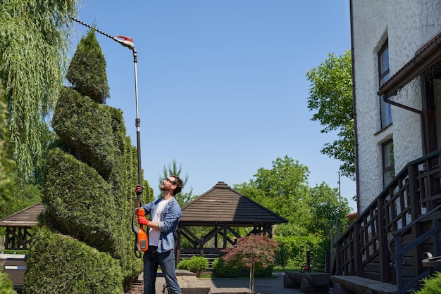 Gratis foto bekwaam tuinier met behulp van een hooggelegen hegtrimmer tijdens het vormgeven van struiken in een topiary-tuin