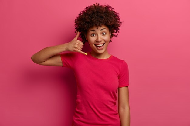 Bel me later. Blij dat een donkere jonge vrouw een telefoongebaar maakt, er positief uitziet met een aantrekkelijke uitdrukking, een brede glimlach, gekleed in vrijetijdskleding, geïsoleerd over een roze muur. Lichaamstaal