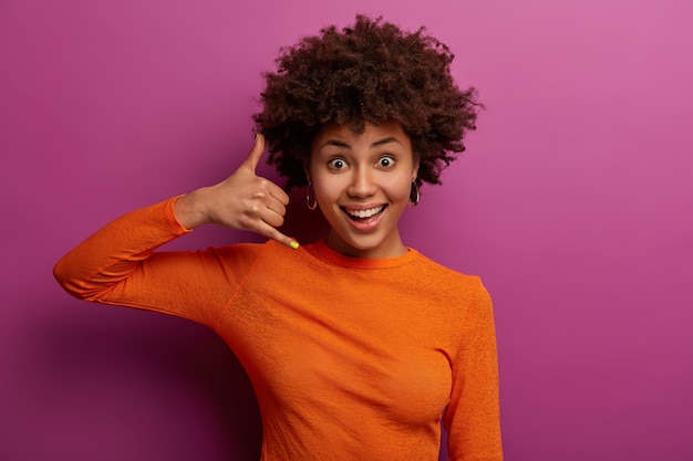 Bel me later. Tevreden gekrulde jonge vrouw maakt telefoongebaar, doet alsof mobiele communicatie, glimlacht gelukkig, draagt casual oranje trui, geïsoleerd over paarse muur. Lichaamstaal concept